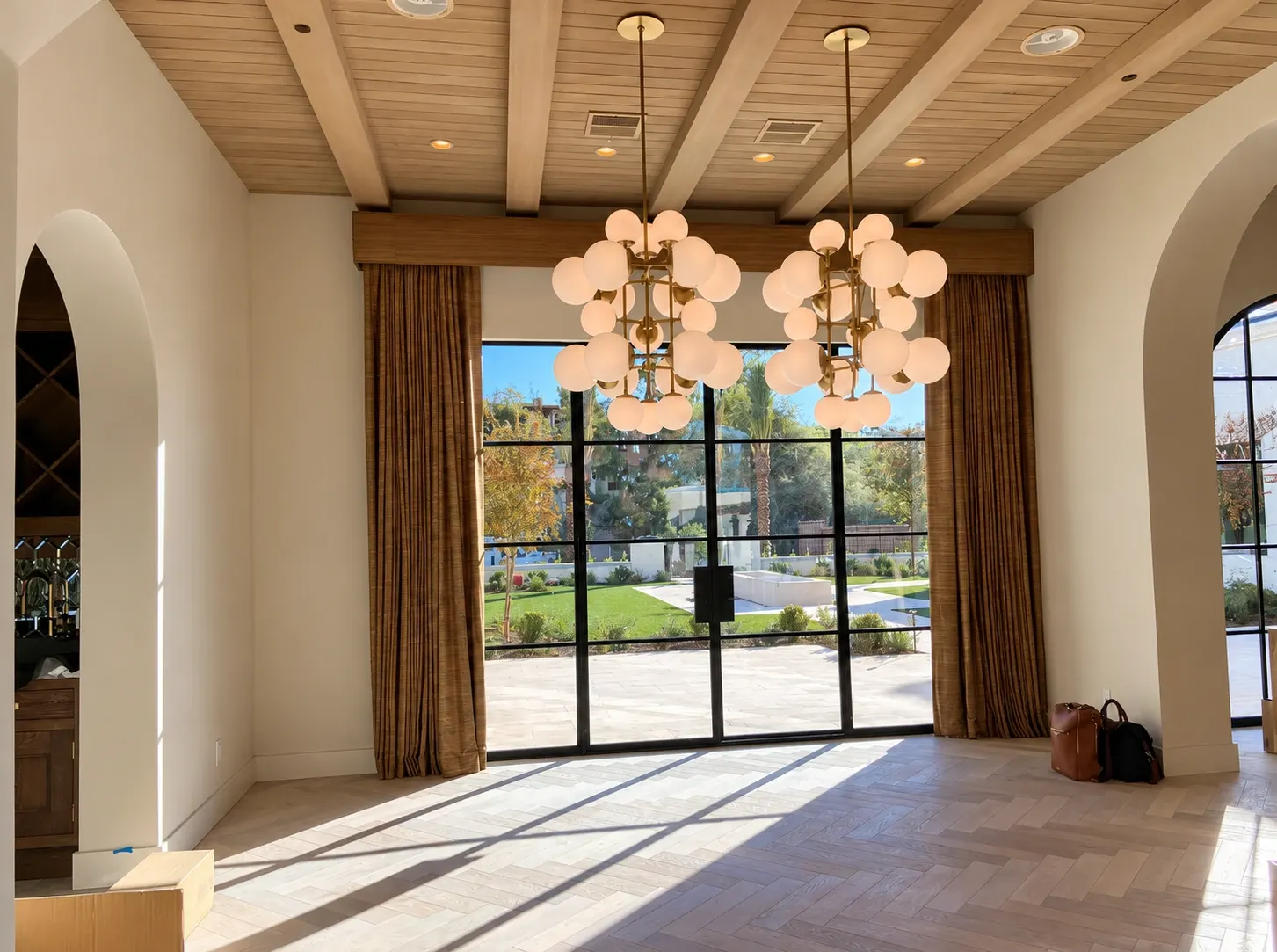 Luxurious modern interior with two pendant chandeliers, wooden ceiling beams, and floor-to-ceiling windows overlooking manicured grounds